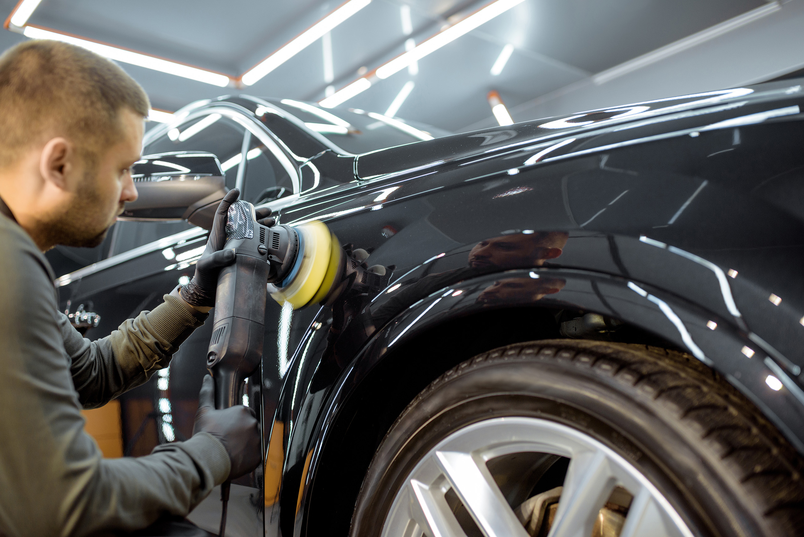 Worker polishing vehicle body with special grinder and wax from scratches at the car service station. Professional car detailing and maintenance concept